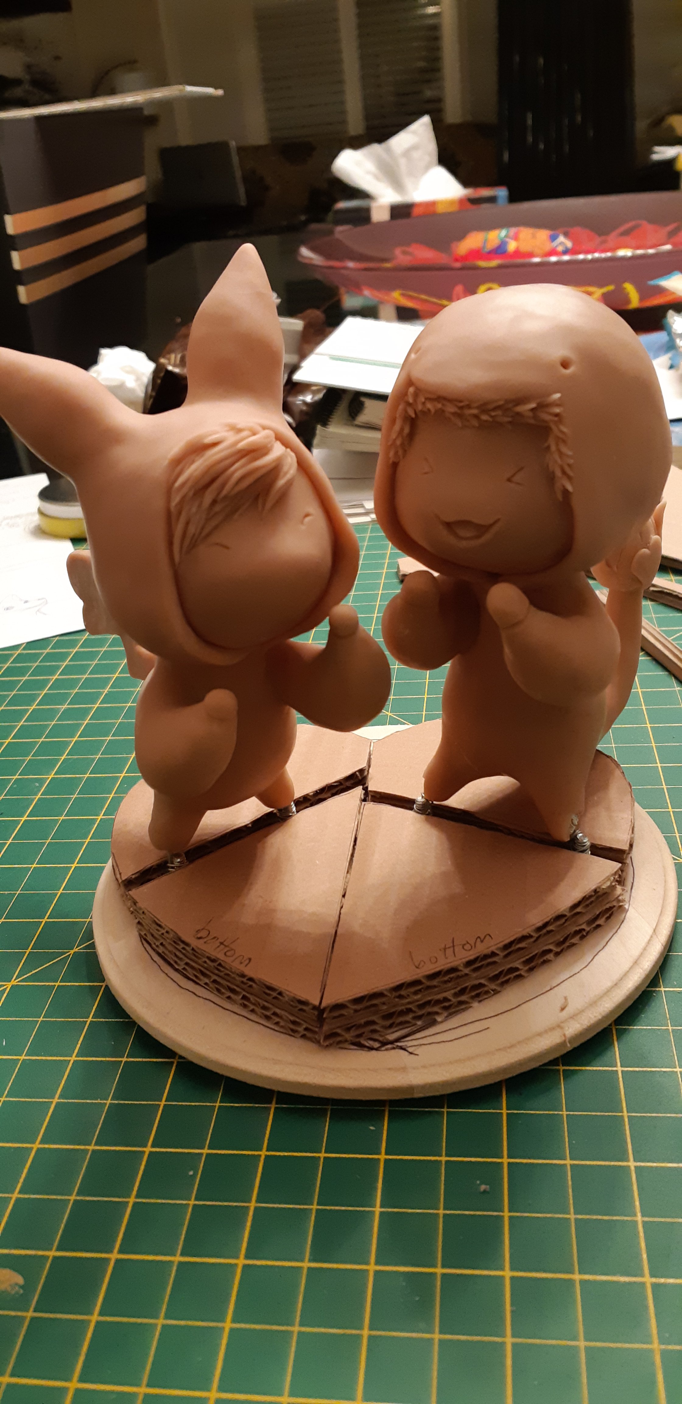 A work-in-progress photo, depicting the unpainted sculpts of the couple. The bodies are covered with sculpey and resemble the couple in the ink drawing. They are fastened to the base and have 4 cardboard segments that assemble to form a heart platform.