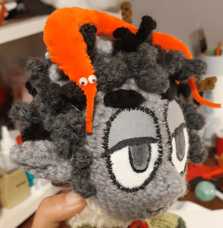 A crocheted Dungeons and Dragons character, named Borgo. The character is a rock gnome, resembling a human-like person with grey skin and pointed ears. Hair is a combination of black and white and is somewhat messy with two twigs sticking out. Character has a bright orange worm-on-a-string sitting on top of his head.
