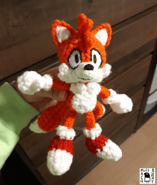 Image of a crocheted fox, named Tails. The character is an orange fox with three orange hairs in the front, white inner ears, muzzle, and tail tips. The character is also wearing white gloves and red shoes with white tips. The crocheted doll is being held to appear standing.