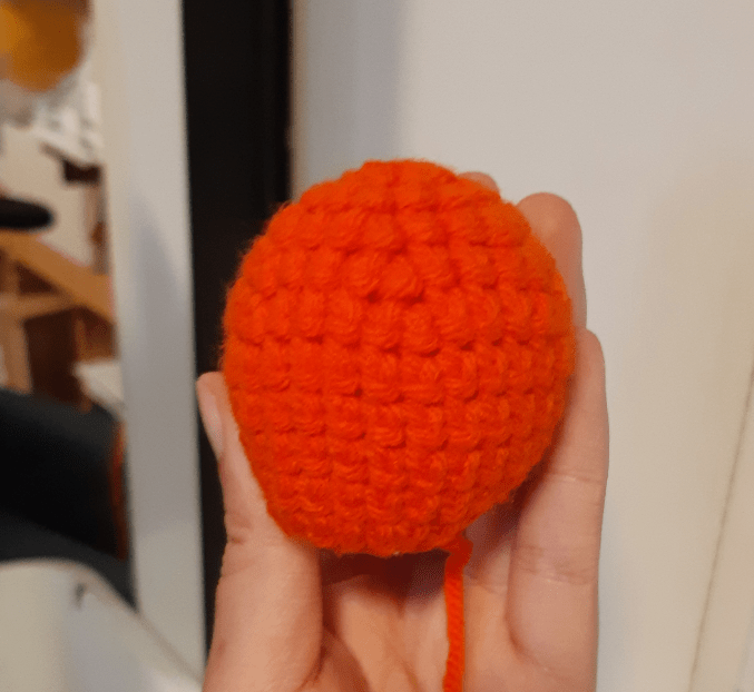 Image of the base part of the crochet doll's head. The shape is somewhat ovoid and the colour is bright orange.