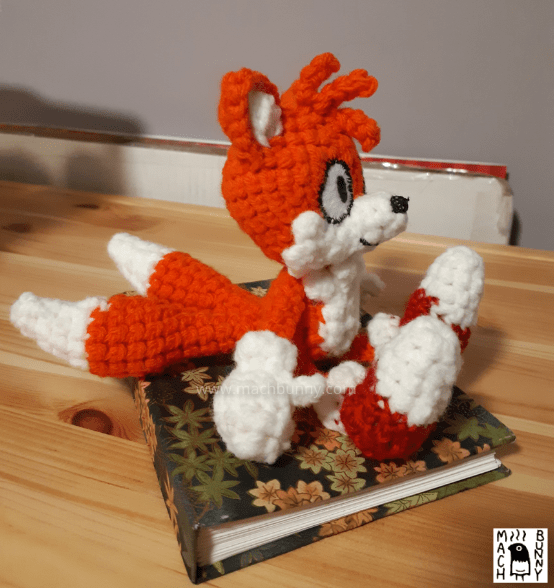 Image of a crocheted fox, named Tails. The character is an orange fox with three orange hairs in the front, white inner ears, muzzle, and tail tips. The character is also wearing white gloves and red shoes with white tips. The crocheted doll is sitting on a notebook. It is a side profile looking towards the right.