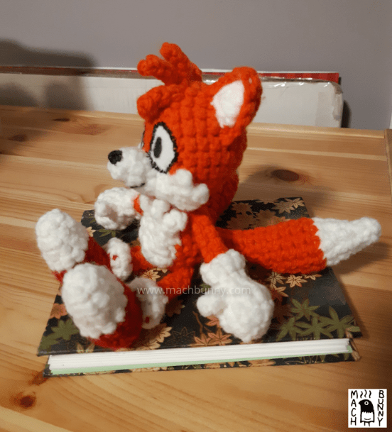 Image of a crocheted fox, named Tails. The character is an orange fox with three orange hairs in the front, white inner ears, muzzle, and tail tips. The character is also wearing white gloves and red shoes with white tips. The crocheted doll is sitting on a notebook. Image is the side profile looking towards the left.