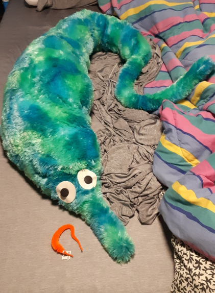 A size comparison between the original worm on a string toy and a giant plush version. The plush version is a teal to blue colour, with eyes made of pleather and buttons.