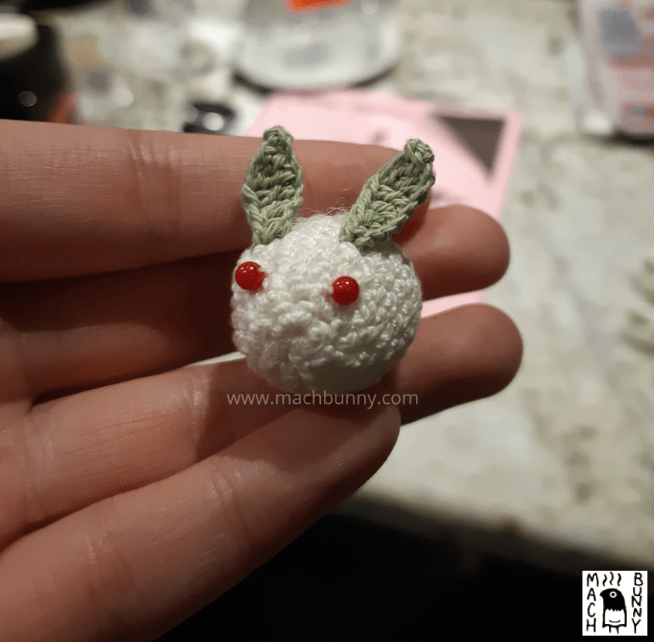 A completed snow bunny, made out of one white oval, two green leaf bits, and two red glass beads. It is facing forward to the left.