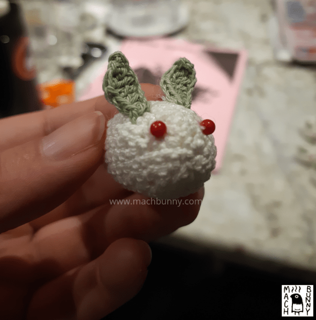 A completed snow bunny, made out of one white oval, two green leaf bits, and two red glass beads. It is facing forward to the right.