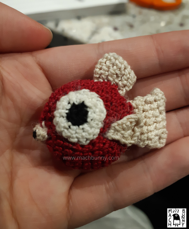 A small crocheted Magikarp, based off of the menu sprites from pokemon gold, silver, and crystal. The piece resembles a small, round, red fish, with large eyes, small lips, and tan-coloured fins. It is smaller than the person's three fingers, where it rests on top.