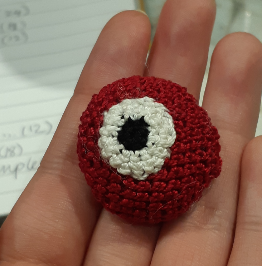 A small crocheted Magikarp, based off of the menu sprites from pokemon gold, silver, and crystal. The piece currently only has the body and eyes put together. It is smaller than the person's three fingers, where it rests on top.