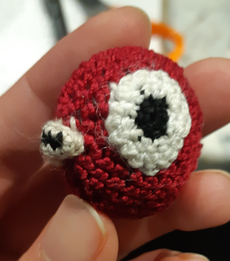 A small crocheted Magikarp, based off of the menu sprites from pokemon gold, silver, and crystal. The piece currently only has the body, eyes, and lips put together. It is smaller than the person's three fingers, where it is being held.