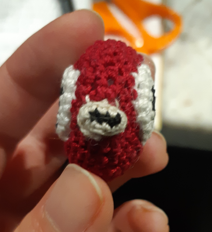 A small crocheted Magikarp, based off of the menu sprites from pokemon gold, silver, and crystal. The piece currently faces forward, and only has the body, eyes, and lips put together. It is smaller than the person's three fingers, where it is being held.