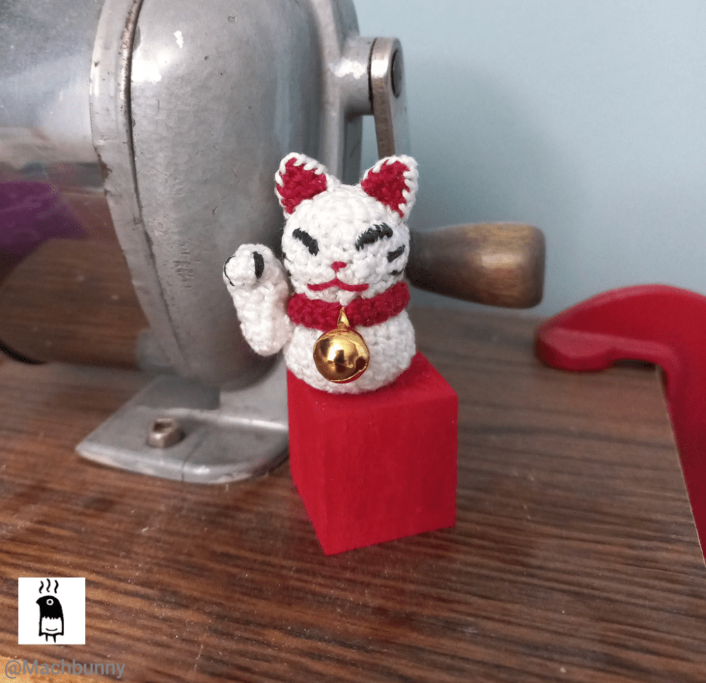 A small crocheted Maneki Neko (Lucky cat), resting on top of a small red wooden block, next to a crank-style pencil sharpener. The cat has one arm in a raised position, and is wearing a red collar with a tiny bell.