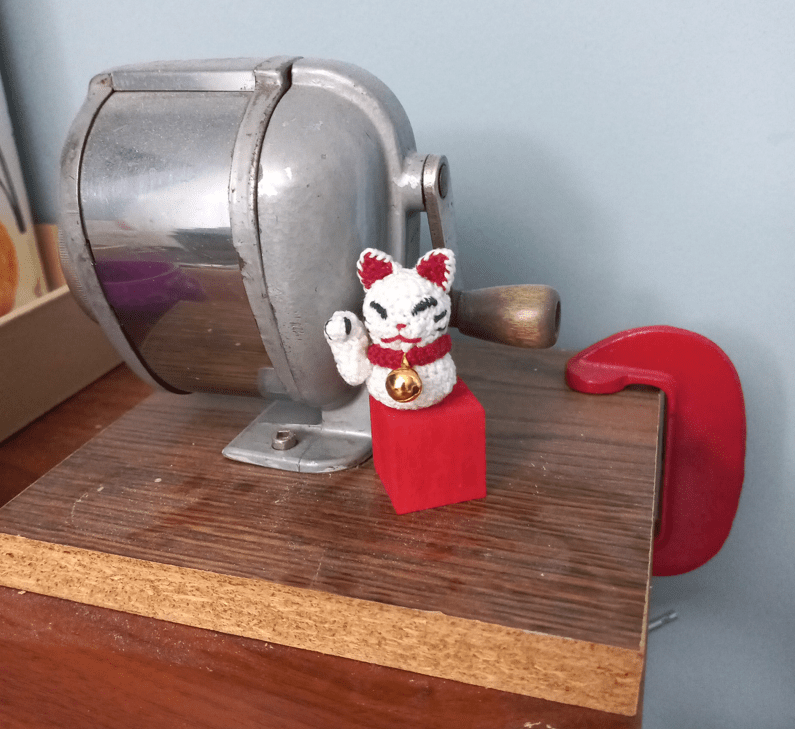 A small crocheted Maneki Neko (Lucky cat), resting on top of a small red wooden block, next to a crank-style pencil sharpener. The cat has one arm in a raised position, and is wearing a red collar with a tiny bell. The angle is slightly further from the cat.