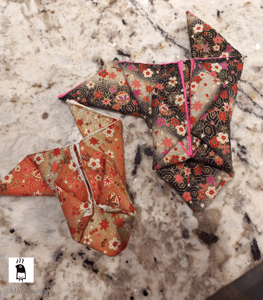 Two origami ballon-style fish, made out of Japanese printed fabric; one is smaller and made with an orange fabric, while the other is made with a black fabric. Both fish are sitting on top of a counter, with the snaps opened and facing down to reveal the undersides. Some of the stitches outline the areas that represent the pouch edges.